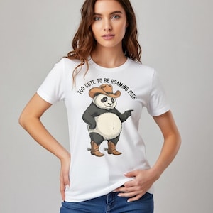 Könnte beinhalten: Weißes T-Shirt mit einem Cartoon-Panda, der einen Cowboyhut und Stiefel trägt. Der Panda zeigt nach rechts. Der Text auf dem Shirt lautet "TOO CUTE TO BE ROAMING FREE".