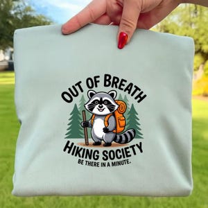 Könnte beinhalten: Hellgrünes T-Shirt mit einem Cartoon-Waschbären, der einen Rucksack trägt und einen Wanderstock hält. Das Design beinhaltet einen Waldhintergrund. Der Text lautet "OUT OF BREATH HIKING SOCIETY BE THERE IN A MINUTE."