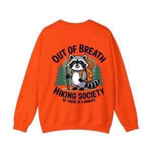 Könnte beinhalten: Orangefarbenes Sweatshirt mit einer Grafik eines Waschbären mit Rucksack und Wanderstock. Der Text lautet "OUT OF BREATH HIKING SOCIETY BE THERE IN A MINUTE."
