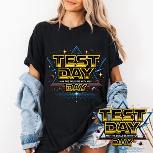 May include: Black t-shirt with a vibrant graphic design. The design features the words "TEST DAY" in a bold, yellow font, with the phrase "MAY THE SKILLS BE WITH YOU" below. The design includes stars, planets, and a geometric background.
