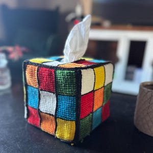 Op de afbeelding: Een kleurrijke tissueboxhoes met een Rubik's Cube-ontwerp. De hoes is gemaakt van stof en heeft een verscheidenheid aan kleuren, waaronder rood, geel, blauw, groen en oranje.