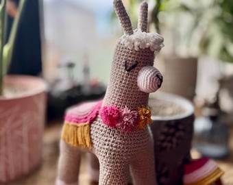 May include: A crocheted llama toy with a pink and yellow saddle and pom-poms. The llama is standing on a wooden surface.