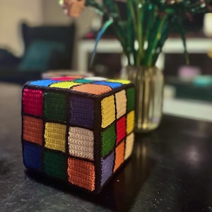 Può includere: Un cubo all'uncinetto a forma di cubo di Rubik con un bordo nero e quadrati colorati. Il cubo è fatto di filati di diversi colori, tra cui rosso, giallo, verde, blu, arancione e bianco.