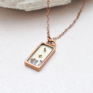 Puede incluir: Un collar de cadena de cobre con un colgante rectangular que contiene pequeñas flores y hojas secas. El colgante está hecho de resina transparente y tiene un marco de cobre. El collar está sobre un fondo blanco.