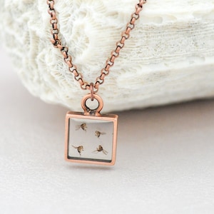 Può includere: Una collana con catena in rame con un ciondolo quadrato che presenta un piccolo quadrato di resina trasparente con fiori secchi all'interno. Il ciondolo è appeso su una superficie bianca.