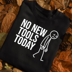 以下が含まれることがあります： 「NO NEW TOOLS TODAY」の白文字がプリントされた黒いクルーネックのスウェットシャツ。テキストの横には、悲しい顔をした白い棒人間が描かれています。スウェットシャツは茶色の葉の上に置かれています。