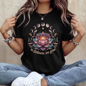 K&ouml;nnte beinhalten: Schwarzes T-Shirt mit einem farbenfrohen Lotusblumen-Design, Mondphasen und dem Text "In Stillness, we grow". Das florale Design umfasst Farbt&ouml;ne von Rosa, T&uuml;rkis und Lila. Das T-Shirt hat einen Rundhalsausschnitt.