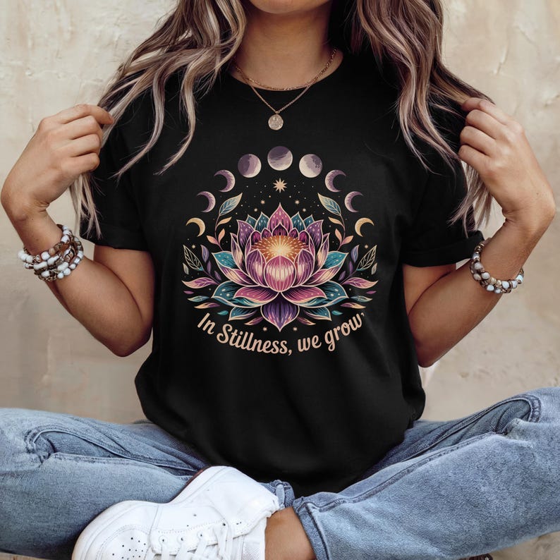 K&ouml;nnte beinhalten: Schwarzes T-Shirt mit einem farbenfrohen Lotusblumen-Design, Mondphasen und dem Text "In Stillness, we grow". Das florale Design umfasst Farbt&ouml;ne von Rosa, T&uuml;rkis und Lila. Das T-Shirt hat einen Rundhalsausschnitt.