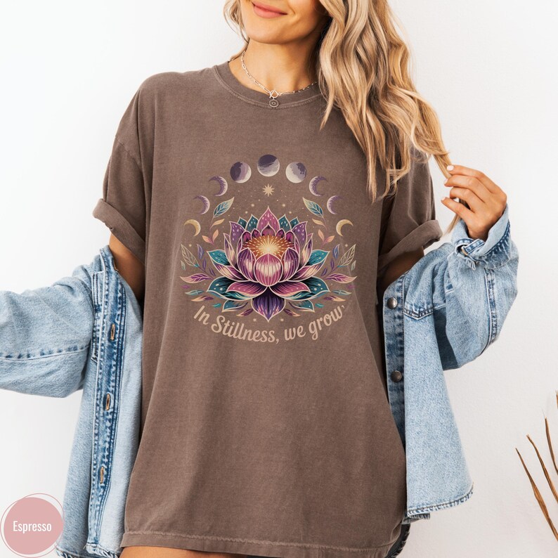 K&ouml;nnte beinhalten: Espressofarbenes T-Shirt mit einem Lotusblumen-Design, Mondphasen und dem Text "In Stillness, we grow". Die Grafik verwendet Lila, Rosa, T&uuml;rkis und Gold. Das Shirt hat hochgekrempelte &Auml;rmel.