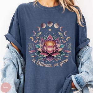 K&ouml;nnte beinhalten: Blaues T-Shirt mit einem Lotusblumen-Design, Mondphasen und dem Text "In Stillness, we grow". Das florale Design umfasst rosa, lila und gr&uuml;ne Farben. Das T-Shirt hat kurze &Auml;rmel und eine lockere Passform.