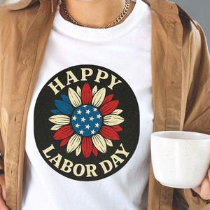 Könnte beinhalten: Weißes T-Shirt mit einem kreisförmigen Aufdruck mit der Aufschrift "HAPPY LABOR DAY". Die Grafik ist ein stilisiertes Blumenmuster mit roten, weißen und blauen Blütenblättern und einem blauen Zentrum mit weißen Sternen. Das T-Shirt wird von einer Person getragen.