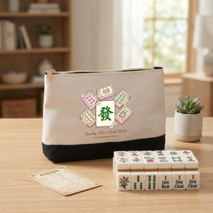 May include: A Mahjong set with a beige and black zippered pouch featuring a design of Mahjong tiles and the text "Lucky Tiles Club 2026". The set includes a stack of tiles and a small card, all on a wooden surface.