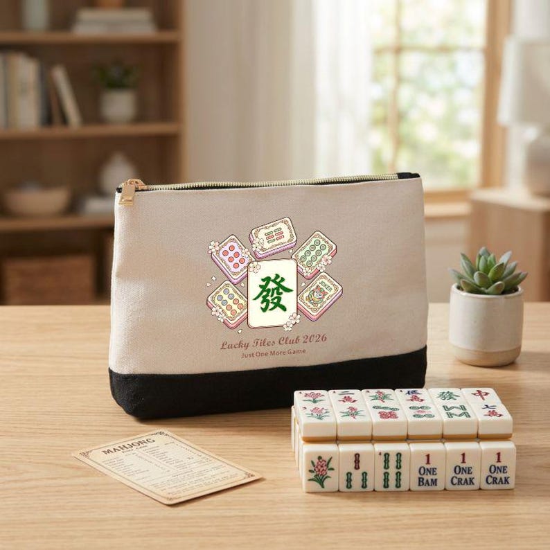 May include: A Mahjong set with a beige and black zippered pouch featuring a design of Mahjong tiles and the text "Lucky Tiles Club 2026". The set includes a stack of tiles and a small card, all on a wooden surface.