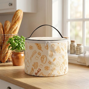 Può includere: Una borsa cilindrica in tessuto con base bianca e un motivo ripetuto a linee marroni raffigurante vari tipi di pane, spighe di grano e la parola BAG. La borsa è dotata di una maniglia nera, una chiusura a zip bianca e un coperchio piatto.