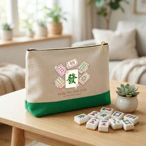 Op de afbeelding: Een beige canvas etui met ritssluiting en een groene basis, met een afbeelding van mahjongtegels en de tekst "Lucky Tiles Club 2026 Just One More Game". Mahjongtegels zijn gerangschikt op een houten tafel.
