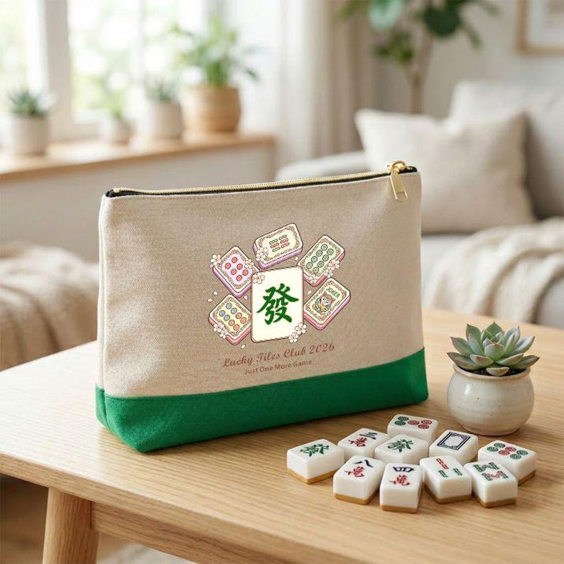 May include: A beige canvas zippered pouch with a green base, featuring a graphic of mahjong tiles and the text "Lucky Tiles Club 2026 Just One More Game." Mahjong tiles are arranged on a wooden table.