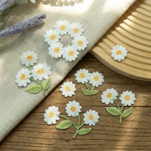 May include: A collection of embroidered daisy patches featuring white petals and yellow centers. The set includes a circular arrangement of six daisies, several individual flower heads, and clusters of two or three daisies with green leaves.