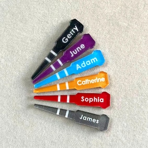 Op de afbeelding: Een verzameling kleurrijke gepersonaliseerde golftees. Elke tee heeft een andere naam: Gerry, June, Adam, Catherine, Sophia en James. De tees zijn zwart, paars, blauw, oranje, rood en grijs met witte strepen.