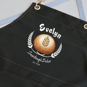 May include: Black apron with the name "Evelyn" in white script above a circular bread design. The design includes a wheat wreath and the words "Sourdough Baker" and "Est. 2026". The apron has a leather strap and metal grommets.