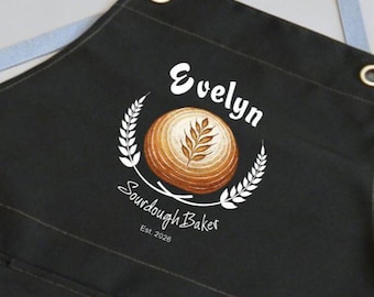 May include: Black apron featuring the name "Evelyn" in white script above a bread design. The design includes a wheat wreath and the words "Sourdough Baker" and "Est. 2026". The apron has a leather strap and metal eyelets.