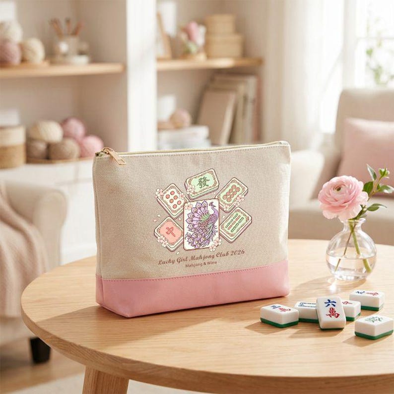 May include: A beige and pink zippered pouch with a Mahjong tile design and the text "Lucky Girl Mahjong Club 2026". The pouch is on a wooden table with Mahjong tiles scattered around it. A small vase with a pink flower is also on the table.