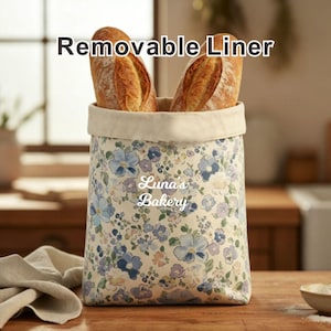 May include: A floral fabric bread basket with a removable liner, holding two baguettes. The basket features a cream-colored rim and a pattern of blue and purple flowers on a cream background. Text on the basket reads "Luna's Bakery". The text "Removable Liner" is above the basket.