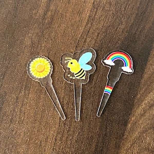 Op de afbeelding: Drie heldere acryl drankroerstaafjes met decoratieve toppers. Eén heeft een gele zonnebloem, een ander een cartoonbij met blauwe vleugels en de derde een regenboog. De roerstaafjes liggen op een bruin oppervlak.