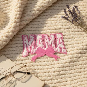 May include: A pink and white floral "MAMA" patch with a pink bow. The patch is on a cream-colored, textured blanket. A sprig of lavender and a pair of gold-rimmed glasses on a book are also in the image.