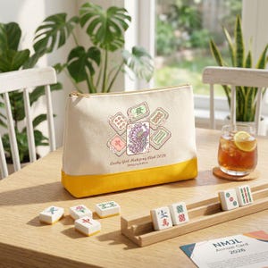 May include: A beige and yellow cosmetic bag with a gold zipper, featuring a peacock and Mahjong tile design. The text "Lucky Girl Mahjong Club 2026" is printed on the bag. Several Mahjong tiles are arranged on a wooden tray.