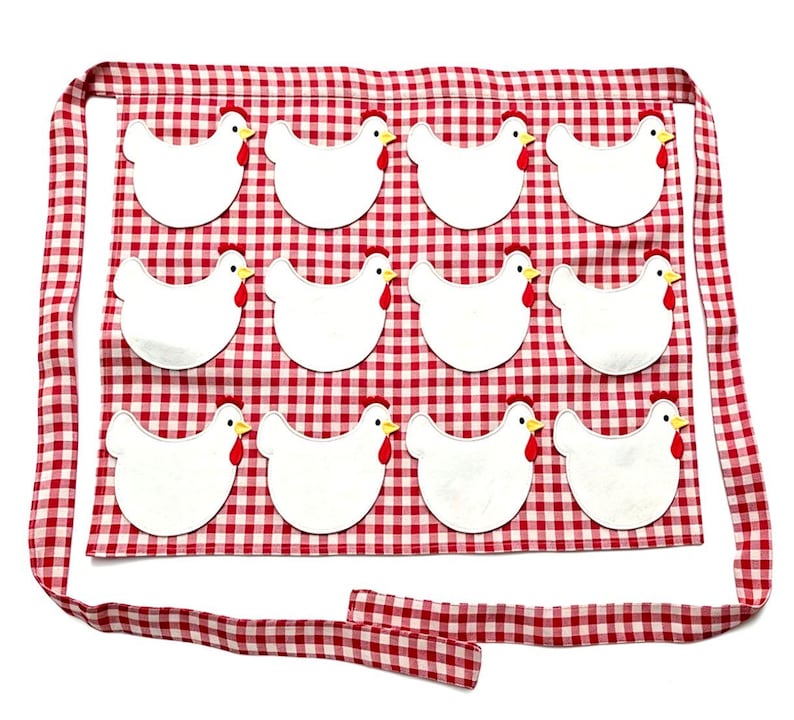 May include: A red and white gingham apron featuring twelve white chicken appliqu&eacute;s arranged in three rows of four. Each chicken has a yellow beak and a red comb. The apron includes long fabric ties at the top and bottom for securing around the waist.