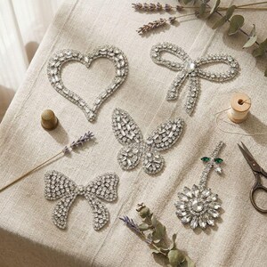 May include: A collection of silver rhinestone embellishments in the shapes of a heart, two bows, a butterfly, and a flower. The image also includes a thimble, lavender sprigs, a spool of thread, and a pair of scissors.