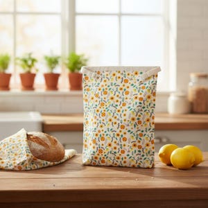 May include: A rectangular bread bag and a matching bread wrap, both adorned with a yellow sunflower pattern on a cream background. The bag has a beige top with a strap. A loaf of bread is wrapped in the matching fabric. Three lemons sit on a wooden surface.