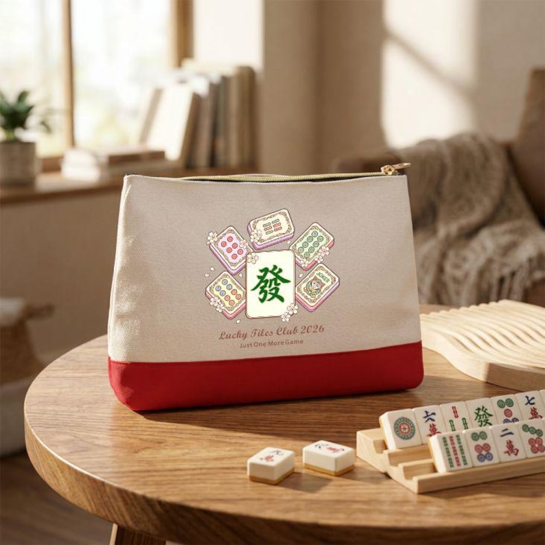 May include: A beige canvas pouch with a red bottom and gold zipper. The pouch features a graphic of mahjong tiles and the text "Lucky Tiles Club 2026 Just One More Game". Mahjong tiles are on a wooden table.