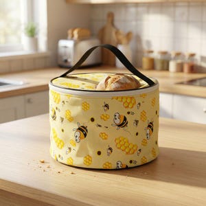 Pode incluir: Um aquecedor de pão redondo de tecido amarelo com alça preta e fecho de zíper. O exterior apresenta um padrão repetitivo de abelhas de desenho animado e favos de mel dourados. Um pão crocante é visível no topo aberto do recipiente.