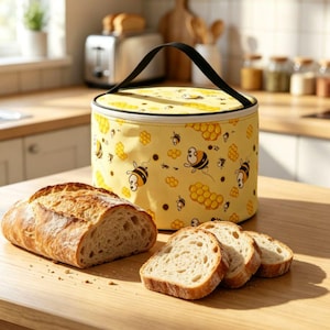 Könnte beinhalten: Ein zylindrischer gelber Brotbeutel mit schwarzem Griff und Reißverschluss, verziert mit einem wiederkehrenden Muster aus Cartoon-Bienen und Waben. Ein knuspriger Brotlaib und drei Brotscheiben liegen vor dem Beutel.