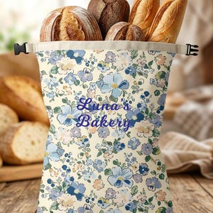 Peut inclure: Un sac à pain en tissu couleur crème orné d'un motif floral bleu et violet avec le texte "Luna's Bakery" imprimé en bleu. Le sac possède une fermeture à enroulement sécurisée par une boucle en plastique noir et contient plusieurs pains.