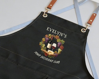 May include: Black apron with a wine-themed design. The apron features a wine bottle, a glass of wine, and a cheese board within a wreath of grapes and leaves. The text "Evelyn's Pour Decisions Club" is printed on the apron.