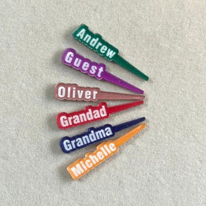 May include: A collection of colorful, personalized name tags. Each tag features a different name: Andrew, Guest, Oliver, Grandad, Grandma, and Michelle. The tags are in various colors, including green, purple, red, blue, and orange.