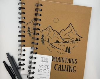 May include: Two spiral-bound notebooks with kraft paper covers. One notebook features a mountain range illustration and the words "MOUNTAINS CALLING." The other notebook has a smaller tag that reads "NOTE BOOK" and "100 pages made in WA state." Two black pens are also visible.