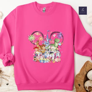 May include: A vibrant pink sweatshirt featuring a colorful graphic of Disney's Toy Story characters. The design includes Woody, Buzz Lightyear, and other beloved characters, set against a whimsical background with palm trees and a castle. The sweatshirt has a crew neck and long sleeves.