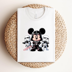 Pu&ograve; includere: T-shirt bianco con un'illustrazione di Mickey Mouse vestito da Darth Vader, affiancato da due Stormtrooper. Mickey Mouse tiene una spada laser rossa. Il disegno &egrave; centrato sulla maglietta.