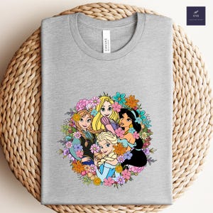 May include: A heather gray t-shirt featuring a colorful graphic of animated princesses surrounded by flowers. The design includes characters from popular animated films. The shirt has a classic crew neck and a tag that reads "CANVAS".