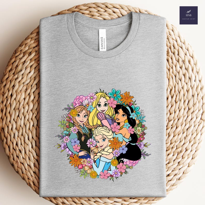 May include: A heather gray t-shirt featuring a colorful graphic of animated princesses surrounded by flowers. The design includes characters from popular animated films. The shirt has a classic crew neck and a tag that reads "CANVAS".