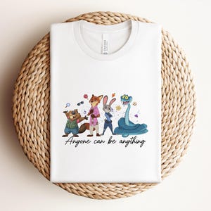 K&ouml;nnte beinhalten: Wei&szlig;es T-Shirt mit Zootopia-Cartoon-Figuren. Das Design zeigt ein Biber, Fuchs, Hase und Schlange, mit dem Text "Anyone can be anything". Das T-Shirt liegt auf einer geflochtenen Oberfl&auml;che.