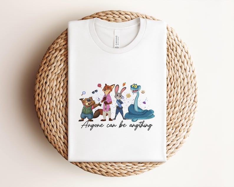 K&ouml;nnte beinhalten: Wei&szlig;es T-Shirt mit Zootopia-Cartoon-Figuren. Das Design zeigt ein Biber, Fuchs, Hase und Schlange, mit dem Text "Anyone can be anything". Das T-Shirt liegt auf einer geflochtenen Oberfl&auml;che.