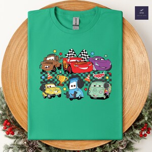 May include: Green t-shirt featuring a colorful graphic of cartoon cars. The design includes characters like Lightning McQueen, Mater, and others, along with checkered flags, stars, and a trophy. The word "Cars" is also visible.