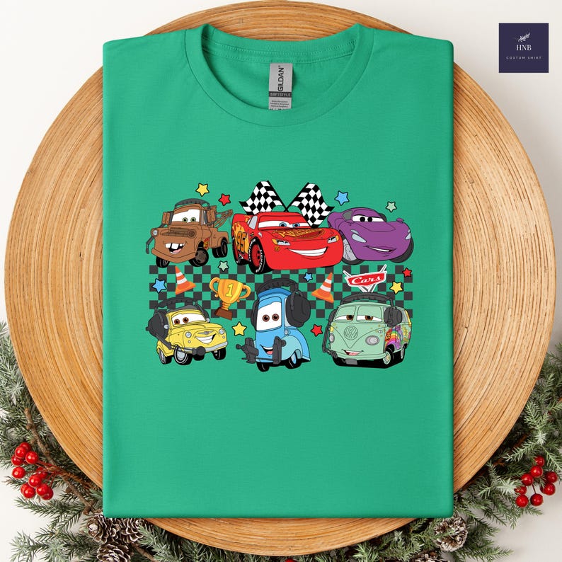 May include: Green t-shirt featuring a colorful graphic of cartoon cars. The design includes characters like Lightning McQueen, Mater, and others, along with checkered flags, stars, and a trophy. The word "Cars" is also visible.