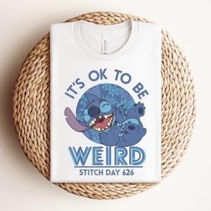 Puede incluir: Camiseta blanca con un personaje de dibujos animados azul y el texto "It's ok to be weird" y "Stitch Day 626". El personaje se r&iacute;e con un ojo cerrado. La camiseta est&aacute; doblada sobre una estera tejida.