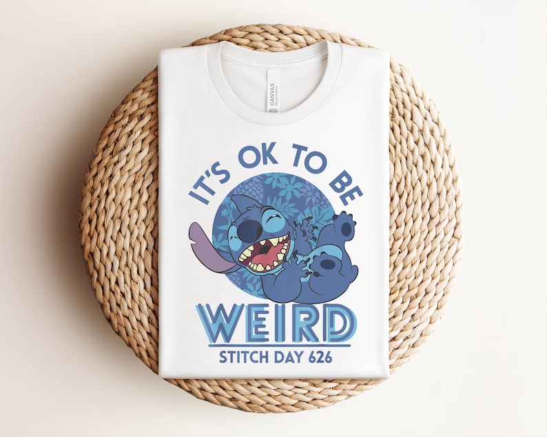 Puede incluir: Camiseta blanca con un personaje de dibujos animados azul y el texto "It's ok to be weird" y "Stitch Day 626". El personaje se r&iacute;e con un ojo cerrado. La camiseta est&aacute; doblada sobre una estera tejida.