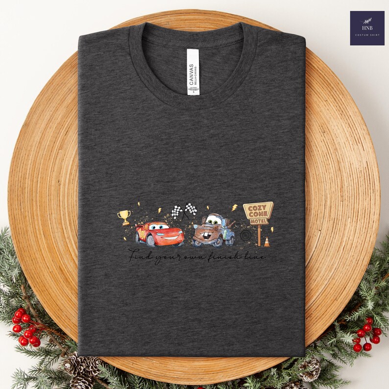 May include: Dark gray t-shirt featuring a graphic of two cartoon cars, a trophy, and checkered flags. The text "Find your own finish line" is below the graphic. The shirt has a crew neck.